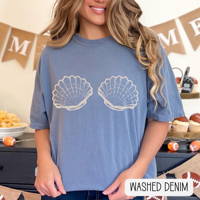 May include: A washed denim blue short-sleeve t-shirt featuring a graphic print of two beige seashells centered on the chest. The shirt has a crew neckline and a relaxed fit. The text "WASHED DENIM" is printed in black in the bottom right corner.