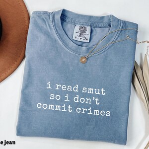 May include: Blue jean-coloured t-shirt with the text "i read smut so i don't commit crimes" in white. The shirt has a crew neck and short sleeves. A gold necklace with a pendant is draped on the shirt.