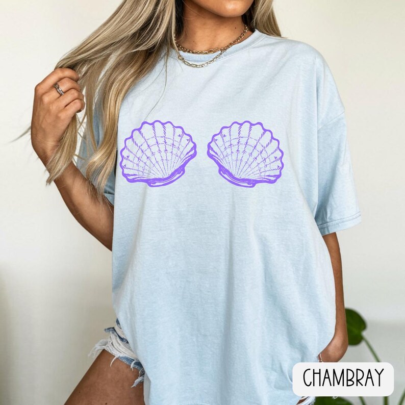May include: A light blue short-sleeved t-shirt featuring a graphic print of two purple seashells positioned side by side on the chest. The shirt has a crew neckline and a relaxed fit, with the word CHAMBRAY printed in black on a white label.