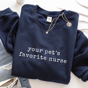 May include: Navy blue sweatshirt with the text "your pet's favorite nurse" in white. A silver necklace with a pendant is draped across the front. The sweatshirt is paired with blue jeans and white shoes.