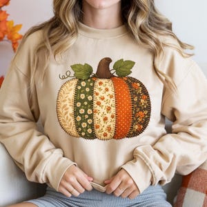 May include: A beige crewneck sweatshirt featuring a large graphic of a pumpkin made from vertical patchwork fabric strips. The pumpkin design includes various floral and polka dot patterns in shades of orange, green, cream, and brown with two green leaves.