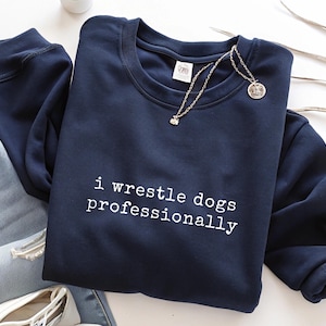 May include: A navy blue crewneck sweatshirt featuring the white text "i wrestle dogs professionally" printed on the front. The garment has long sleeves with ribbed cuffs and a ribbed neckline, paired with light blue denim jeans and a silver necklace.