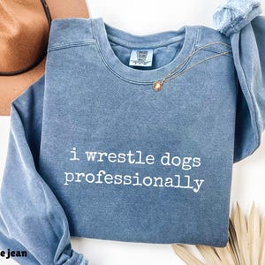 May include: A blue jean-colored sweatshirt with the text "i wrestle dogs professionally" in white. A gold necklace with a pendant is draped on the shirt. A brown hat is in the background.