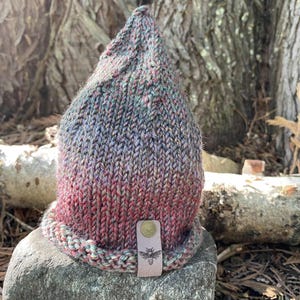 May include: A knitted beanie hat with a pointed crown. The hat has a colour gradient, transitioning from red at the base to blue and green at the top. A leather tag with a bee emblem is attached to the brim.