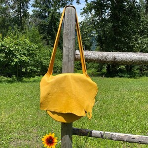 May include: A mustard yellow leather shoulder bag with long straps, hanging on a wooden post. The bag has a rounded bottom and a textured surface. The background includes a green grassy area, trees, and a wooden fence.