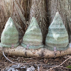 Peut inclure: Trois chapeaux tricotés verts avec des pointes et des bords roulés sont exposés sur une bûche. Chaque chapeau a une petite étiquette en cuir avec un emblème d'abeille. Les chapeaux sont de différentes nuances de vert, certains avec un aspect moucheté.