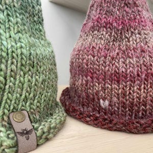 May include: Two knitted beanies. One is green with a leather tag featuring a bee emblem. The other is a mix of red, pink, and brown. Both beanies are made of thick, textured yarn, perfect for cold weather.