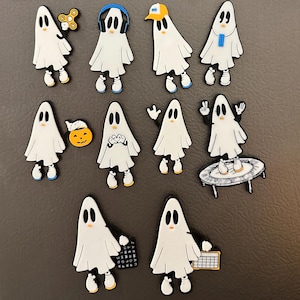 May include: A collection of ten white ghost magnets with various designs. The ghosts have different facial expressions, hats, and accessories, including a fidget spinner, headphones, and a candy corn.
