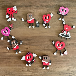 May include: A collection of nine heart-shaped and chocolate bar-shaped magnets with cartoon faces. The magnets are in various colors, including red, pink, brown, and white. The magnets are arranged in a circle on a wooden surface.