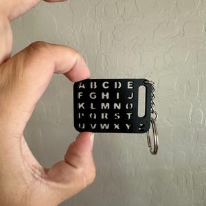 May include: A black keychain with a metal ring and a rectangular tag. The tag has all the letters of the alphabet cut out in a grid pattern.