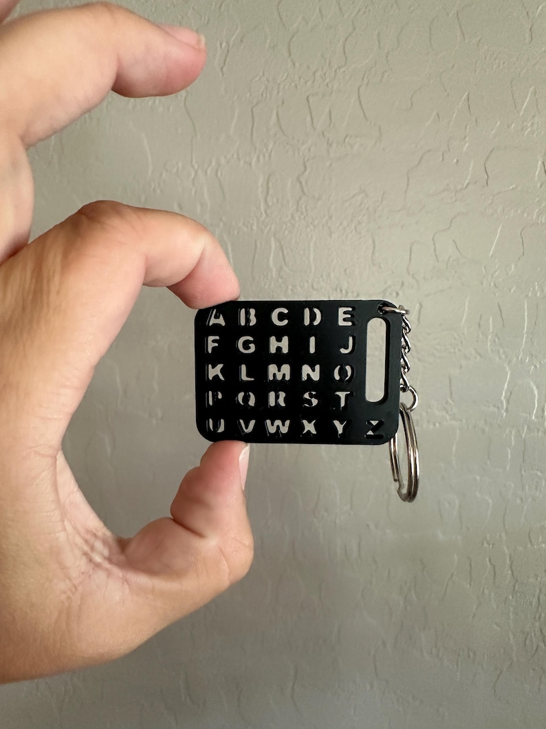 May include: A black keychain with a metal ring and a rectangular tag. The tag has all the letters of the alphabet cut out in a grid pattern.