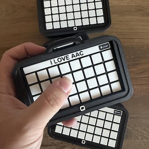 May include: A black rectangular frame with a grid of white square tiles. The top row features the text I LOVE AAC in black letters. The frame includes a small handle at the top and a square button icon at the bottom center of the frame border.