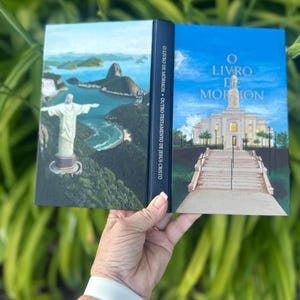 May include: A book with a painted cover showing a landscape of Christ the Redeemer statue and a white temple. The spine reads "O LIVRO DE MORMON". The book is held against a backdrop of green foliage.