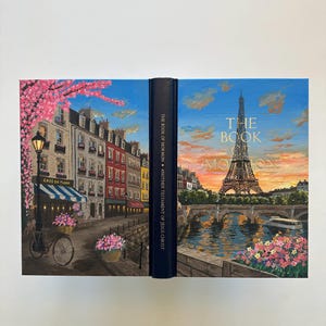 May include: A hardback book titled The Book of Mormon: Another Testament of Jesus Christ. The cover features a painted scene of a Parisian street with a cafe, a bicycle, and the Eiffel Tower over the Seine river at sunset with pink flowers.
