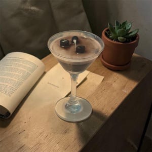 Op de afbeelding: Een helder cocktailglas gevuld met een donkerbruine vloeistof en gegarneerd met drie donkere, ronde voorwerpen. Rechts staat een kleine vetplant in een pot. Een open boek en een stuk papier liggen op het houten oppervlak.