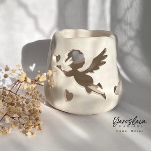 Peut inclure: Bougeoir en céramique blanc cassé avec un motif d'ange découpé, des cœurs et une ouverture carrée. Le bougeoir a une finition mate et convient à la décoration intérieure. Le texte "Yaroslav Mariani Home Décor" est en bas à droite.