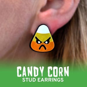May include: A close-up of a candy corn stud earring. The earring is shaped like a candy corn with a white top, yellow centre, and orange base. The candy corn has an angry face with black eyes and a frown. The text "CANDY CORN STUD EARRINGS" is at the bottom.