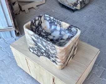 May include: A rectangular marble sink with a speckled pattern of white, black, and tan. The sink is set on a wooden box. The interior of the sink has a smooth, polished finish, and a drain hole is visible.