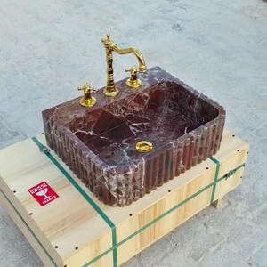 K&ouml;nnte beinhalten: Ein rechteckiges Waschbecken aus rotem Marmor mit goldener Armatur und Armaturen. Das Waschbecken steht auf einer Holzkiste mit gr&uuml;nen Gurten und einem roten "Zerbrechlich"-Aufkleber. Das Innere des Waschbeckens hat ein Muster.