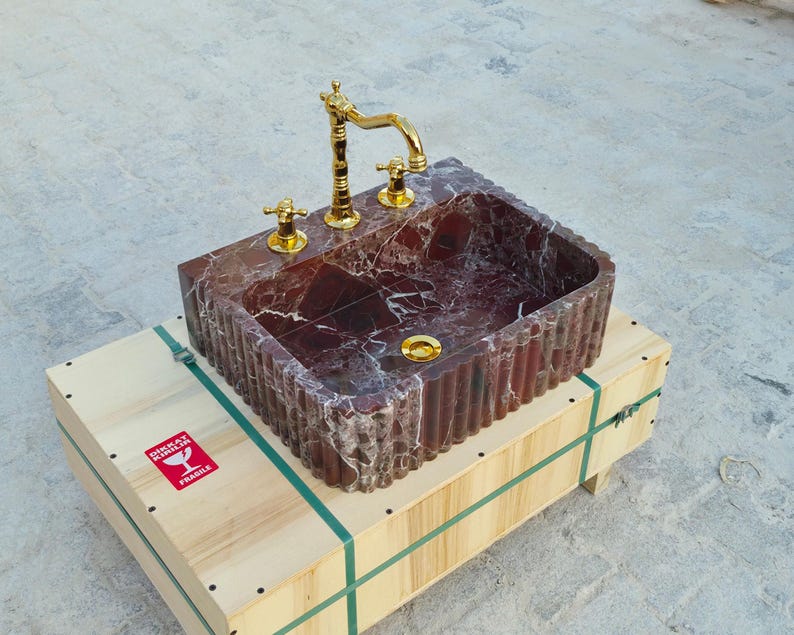 K&ouml;nnte beinhalten: Ein rechteckiges Waschbecken aus rotem Marmor mit goldener Armatur und Armaturen. Das Waschbecken steht auf einer Holzkiste mit gr&uuml;nen Gurten und einem roten "Zerbrechlich"-Aufkleber. Das Innere des Waschbeckens hat ein Muster.