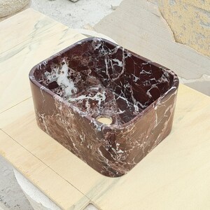 May include: A rectangular bathroom sink with rounded corners. The sink is made of dark red marble with white veining. It has a single drain hole and is set on a light-colored wooden surface.