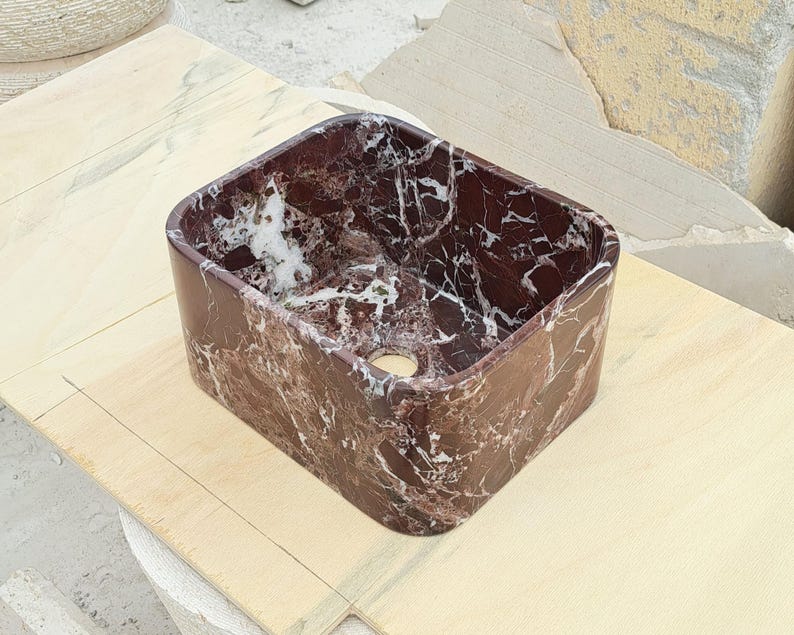 May include: A rectangular bathroom sink with rounded corners. The sink is made of dark red marble with white veining. It has a single drain hole and is set on a light-colored wooden surface.