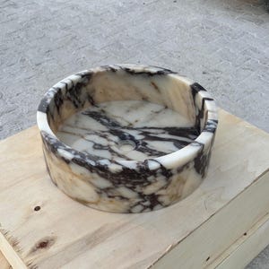 以下が含まれることがあります： ベージュと濃い茶色の模様が入った丸い大理石の洗面台。洗面台は円筒形で、中央に排水穴があります。明るい色の木製の表面に置かれています。
