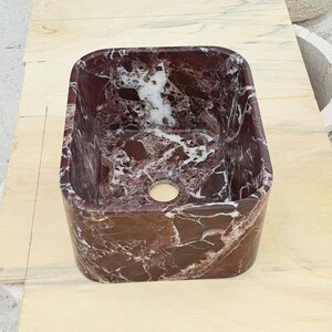 May include: A rectangular, burgundy marble sink with white veining. The sink has a rounded interior and a circular drain hole. The sink is made of natural stone and is suitable for a bathroom or powder room.