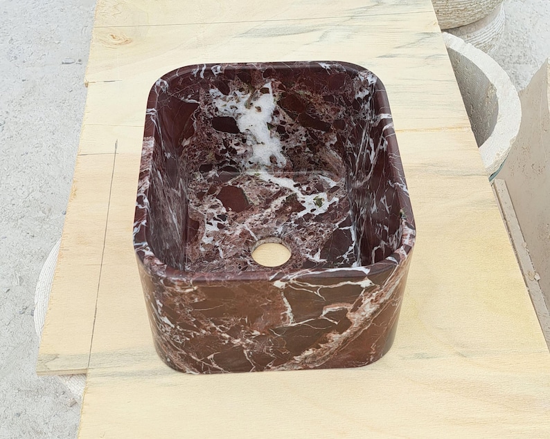 May include: A rectangular, burgundy marble sink with white veining. The sink has a rounded interior and a circular drain hole. The sink is made of natural stone and is suitable for a bathroom or powder room.