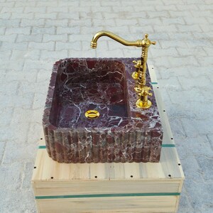 K&ouml;nnte beinhalten: Ein rechteckiges Waschbecken aus burgunderfarbenem Marmor mit goldenen Armaturen und einem goldenen Wasserhahn. Das Waschbecken steht auf einer Holzkiste. Ma&szlig;e in Zentimetern.