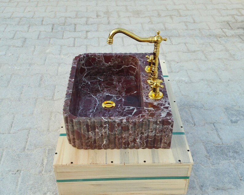K&ouml;nnte beinhalten: Ein rechteckiges Waschbecken aus burgunderfarbenem Marmor mit goldenen Armaturen und einem goldenen Wasserhahn. Das Waschbecken steht auf einer Holzkiste. Ma&szlig;e in Zentimetern.