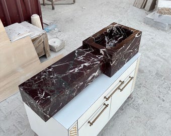 Könnte beinhalten: Ein rechteckiges Waschbecken aus dunkelrotem Marmor mit weißen Adern steht auf einem weißen Schrank mit Messinggriffen. Das Waschbecken hat ein eingebautes Becken und eine lange Thekenverlängerung. Der Marmor hat eine polierte Oberfläche.