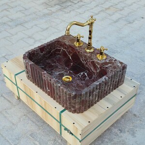 K&ouml;nnte beinhalten: Ein rechteckiges Waschbecken aus rotem Marmor mit einer goldenen Armatur und Armaturen. Das Waschbecken steht auf einer Holzkiste mit gr&uuml;nen Riemen. Der Marmor hat wei&szlig;e Adern und das Waschbecken hat eine gerippte Au&szlig;enseite.
