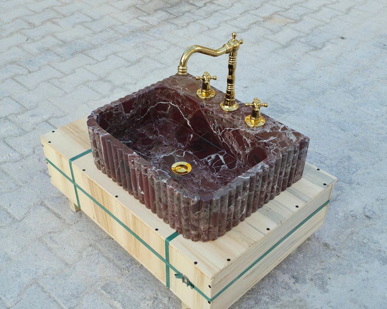 K&ouml;nnte beinhalten: Ein rechteckiges Waschbecken aus rotem Marmor mit einer goldenen Armatur und Armaturen. Das Waschbecken steht auf einer Holzkiste mit gr&uuml;nen Riemen. Der Marmor hat wei&szlig;e Adern und das Waschbecken hat eine gerippte Au&szlig;enseite.