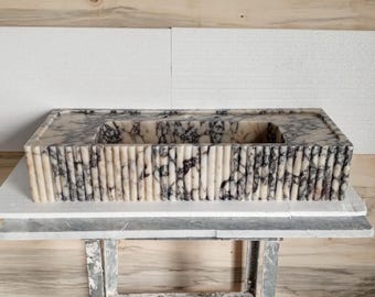Op de afbeelding: Een rechthoekige marmeren wastafel met een geribbelde rand en een wit en paars marmerpatroon. De wastafel staat op een houten tafel.