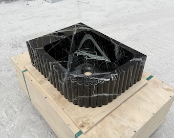 May include: A rectangular black marble sink with white veining. The sink has a fluted exterior design and a central drain hole. It is sitting on a wooden crate.