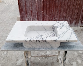 May include: A white marble bathroom sink with a rectangular countertop and a single basin. The sink is sitting on a wooden table.