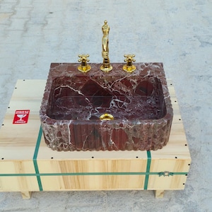 K&ouml;nnte beinhalten: Ein rechteckiges Waschbecken aus rotem Marmor mit goldfarbenen Wasserh&auml;hnen und Armaturen. Das Waschbecken steht auf einer Holzkiste mit gr&uuml;nen Gurten und einem "FRAGILE" Aufkleber.