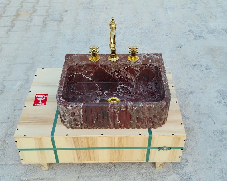 K&ouml;nnte beinhalten: Ein rechteckiges Waschbecken aus rotem Marmor mit goldfarbenen Wasserh&auml;hnen und Armaturen. Das Waschbecken steht auf einer Holzkiste mit gr&uuml;nen Gurten und einem "FRAGILE" Aufkleber.