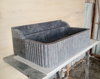 May include: A rectangular, grey stone sink with a fluted front design. The sink has a raised back and is set on a light-coloured surface. The overall design is modern and minimalist, suitable for a bathroom.