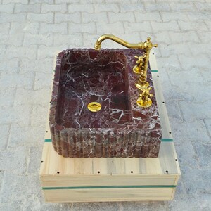 K&ouml;nnte beinhalten: Ein rechteckiges Waschbecken aus burgunderfarbenem Marmor mit wei&szlig;en Adern, mit einer goldfarbenen Armatur und einem Abfluss. Das Waschbecken steht auf einer Holzkiste mit gr&uuml;nen B&auml;ndern. Die Armatur hat ein klassisches Design.