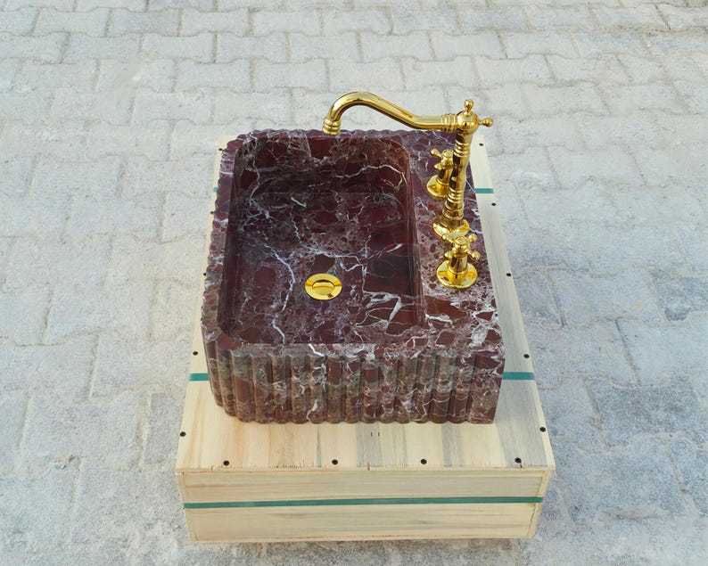 K&ouml;nnte beinhalten: Ein rechteckiges Waschbecken aus burgunderfarbenem Marmor mit wei&szlig;en Adern, mit einer goldfarbenen Armatur und einem Abfluss. Das Waschbecken steht auf einer Holzkiste mit gr&uuml;nen B&auml;ndern. Die Armatur hat ein klassisches Design.