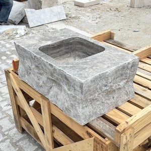 Peut inclure: Un grand évier en pierre grise avec un bassin carré. L'évier a une surface rugueuse et texturée et repose sur une palette en bois. La pierre a un aspect naturel et vieilli, suggérant une utilisation en extérieur. L'évier est probablement destiné à un jardin ou à un espace extérieur.