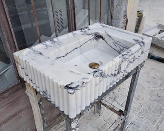 May include: A white marble sink with a fluted edge and a drain in the centre. The sink is on a metal stand.