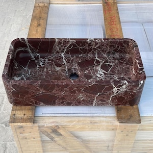 May include: A rectangular, burgundy marble sink with white veining. The sink has rounded corners and a central drain hole. It is displayed on a wooden crate.