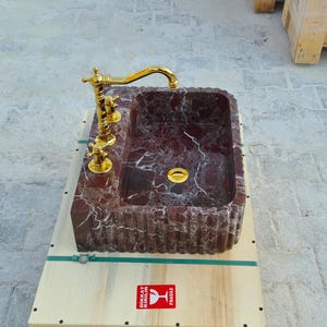 K&ouml;nnte beinhalten: Ein rechteckiges Waschbecken aus Marmor mit einem burgunderroten und wei&szlig;en Adernmuster. Es verf&uuml;gt &uuml;ber eine goldfarbene Armatur und Beschl&auml;ge. Das Waschbecken steht auf einem Holzsockel.