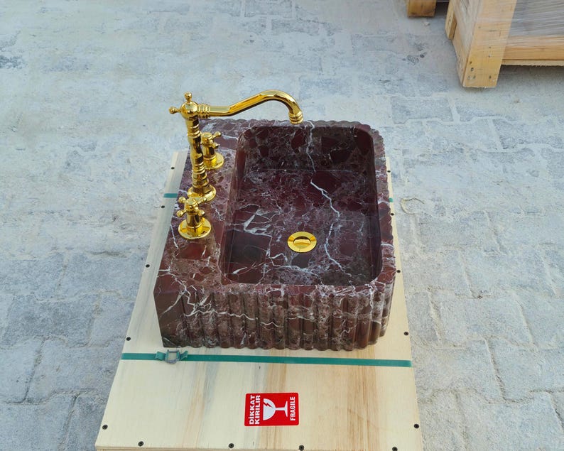 K&ouml;nnte beinhalten: Ein rechteckiges Waschbecken aus Marmor mit einem burgunderroten und wei&szlig;en Adernmuster. Es verf&uuml;gt &uuml;ber eine goldfarbene Armatur und Beschl&auml;ge. Das Waschbecken steht auf einem Holzsockel.