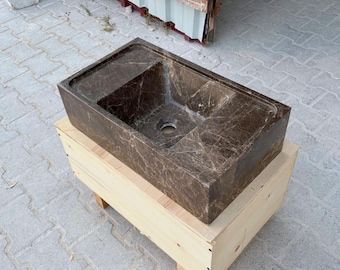 May include: A rectangular brown marble sink with a central drain hole. The sink has a textured surface with light veining. It rests on a light-colored wooden crate, showcasing a natural stone bathroom fixture.