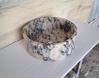 May include: A round white marble sink with black veining. The sink is sitting on a white surface.