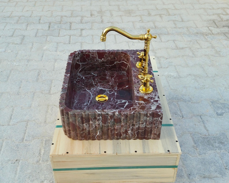 K&ouml;nnte beinhalten: Ein rechteckiges Waschbecken aus Marmor mit einem burgunderroten und wei&szlig;en Adernmuster. Es verf&uuml;gt &uuml;ber einen goldfarbenen Wasserhahn und Armaturen. Das Waschbecken steht auf einer Holzkiste und pr&auml;sentiert eine luxuri&ouml;se Badezimmerausstattung.